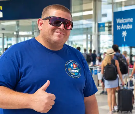 A smiling Dusk till Dawn host in a branded T-shirt gives a thumbs up at the airport, under a Welcome to Aruba sign